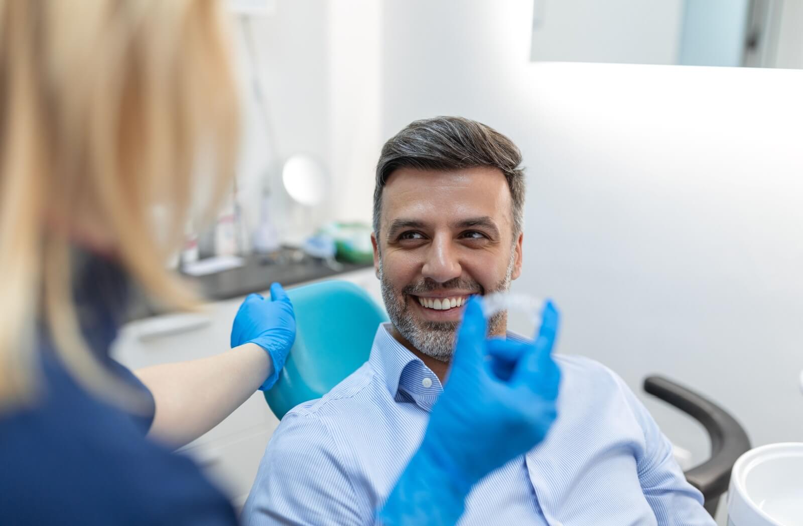 A smiling man sitting in a dental chair while a dental professional, wearing blue gloves, holds up a clear Invisalign aligner during a consultation.